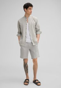 Hellgrau langärmliges Hemd mit Knopfleiste über einem weißen Tanktop, kombiniert mit gestreiften Shorts und schwarzen Sandalen. Besitzt einen lässigen Schnitt und hochgekrempelte Ärmel.