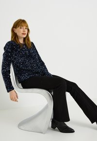 Top de manga larga con lentejuelas azul marino y superficie texturizada, combinado con pantalones negros acampanados y botas negras de tacón, sentado en una silla moderna plateada.