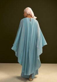 Prenda de chiffon azul claro fluida con diseño de capa ancha, que presenta un detalle de abertura en la espalda y se combina con un pañuelo rosa en la cabeza.