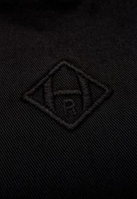 Schwarzer Stoff mit einem erhabenen, schwarzen gestickten Emblem in Diamantenform und geometrischen Designdetails, vor einem strukturierten Hintergrund.