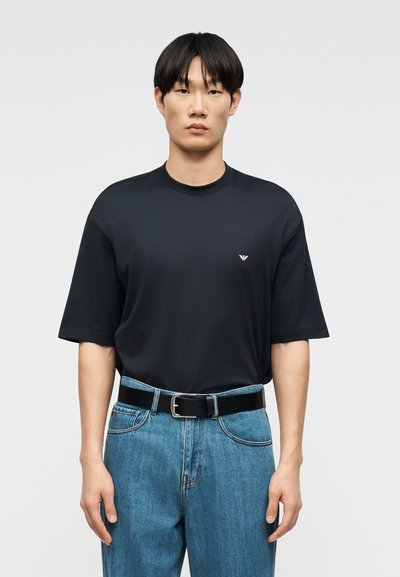 Νέος άνδρας με μαύρα μαλλιά, φορώντας μια απλή μαύρη κοντομάνικη μπλούζα και μπλε τζιν με μαύρη ζώνη, στέκεται μπροστά σε λευκό φόντο.