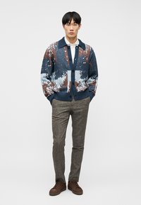 Άνδρας με πολύχρωμο πουλόβερ με γιακά πάνω από λευκό πουκάμισο, γκρι παντελόνι από τσέγκε, και καφέ αθλητικά, στέκεται με τα χέρια στις τσέπες.