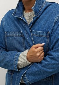 Person i en blå denimjacka som bärs över en beige stickad tröja, med en hand som håller jackan stängd vid bröstet.