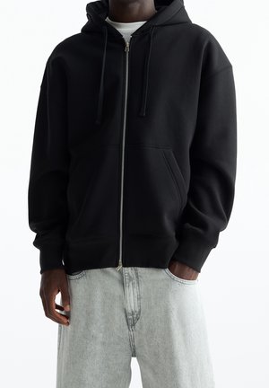 Hombre con sudadera negra con cremallera y cordones, y vaqueros de mezclilla gris claro, con una mano en el bolsillo, sobre un fondo blanco.