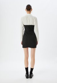 Schwarzes Nadelstreifen-Kleid mit figurbetontem Taillenbereich, kombiniert mit einem weißen Langarmhemd. Besitzt eine glatte Textur und einen geraden Saum.