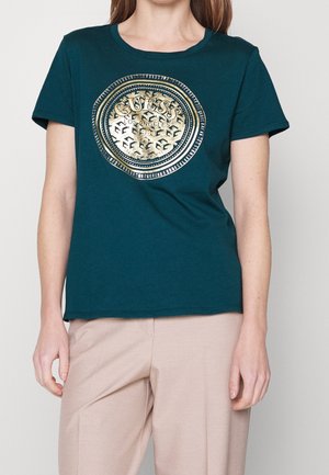 Kobieta ubrana w turkusowy t-shirt z krótkim rękawem z złotym okrągłym emblemtem "GUESS Los Angeles 1981", w połączeniu z beżowymi spodniami.