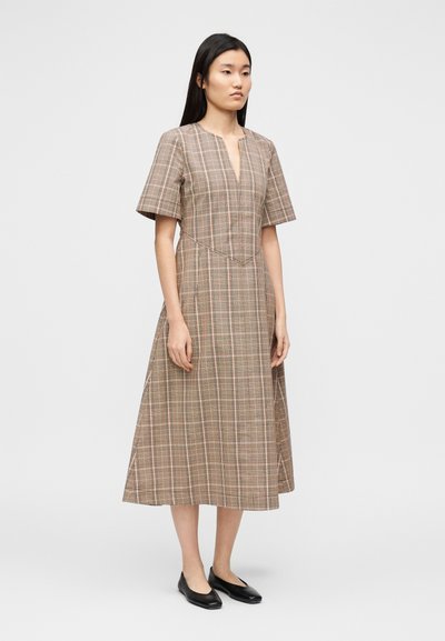 Καφέ καρό φόρεμα με κοντά μανίκια, V-λαιμόκοψη και εφαρμοστό μπούστο. Η φούστα ρέει κάτω από το γόνατο, με ένα διακριτικό φαρδύ τελείωμα.