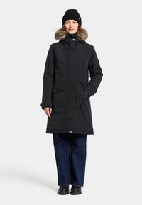 Sort parka med pelsforet hætte, frontlukning med knapper, to sidelommer og et mellemlangt design. Kombination med mørke jeans og sorte støvler.