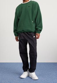 Zelená fleecová mikina se zipovou kapsou, v kombinaci s černými cargo kalhotami a bílými teniskami. Na mikině i kalhotách je logo Nike.