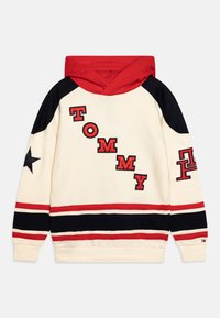 Kermanvärinen huppari punaisilla yksityiskohdilla, mustilla verkkokäsivarsilla ja kirjatuilla "TOMMY"-kirjaimilla. Helmassa mustat ja punaiset raidat.