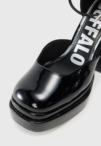 Sapatos de plataforma em pele envernizada preta, com bico arredondado, tira no tornozelo e um salto robusto. Apresentam uma superfície lisa e brilhante, além de uma decoração ousada.