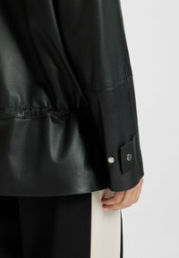 Black leather jacket with a fitted design, featuring a slightly gathered hem and snap button details on the cuffs. Smooth texture overall.