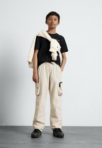 T-shirt noir, sweat-shirt crème drapé sur l'épaule, pantalon cargo beige avec poches, et baskets noires sur un sol gris contre un fond clair.