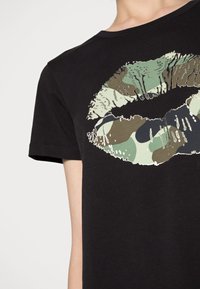 Svart bomullströja med ett stort kysstryck i camouflagefärger, med texturerade detaljer på läpparna och en klassisk rund halsringning.