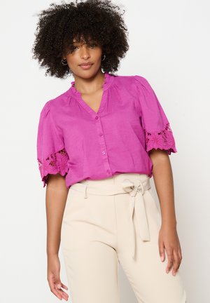 Blusa fucsia con bottoni e scollo arricciato, maniche corte con bordo in pizzo, abbinata a pantaloni beige a vita alta con cintura a cravatta.
