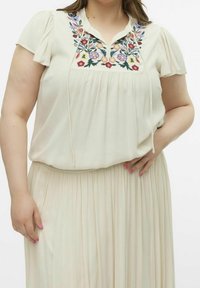 Blouse de couleur crème avec broderies florales au niveau du col, en tissu doux, à manches courtes bouffantes et coupe ample, associée à une jupe plissée.