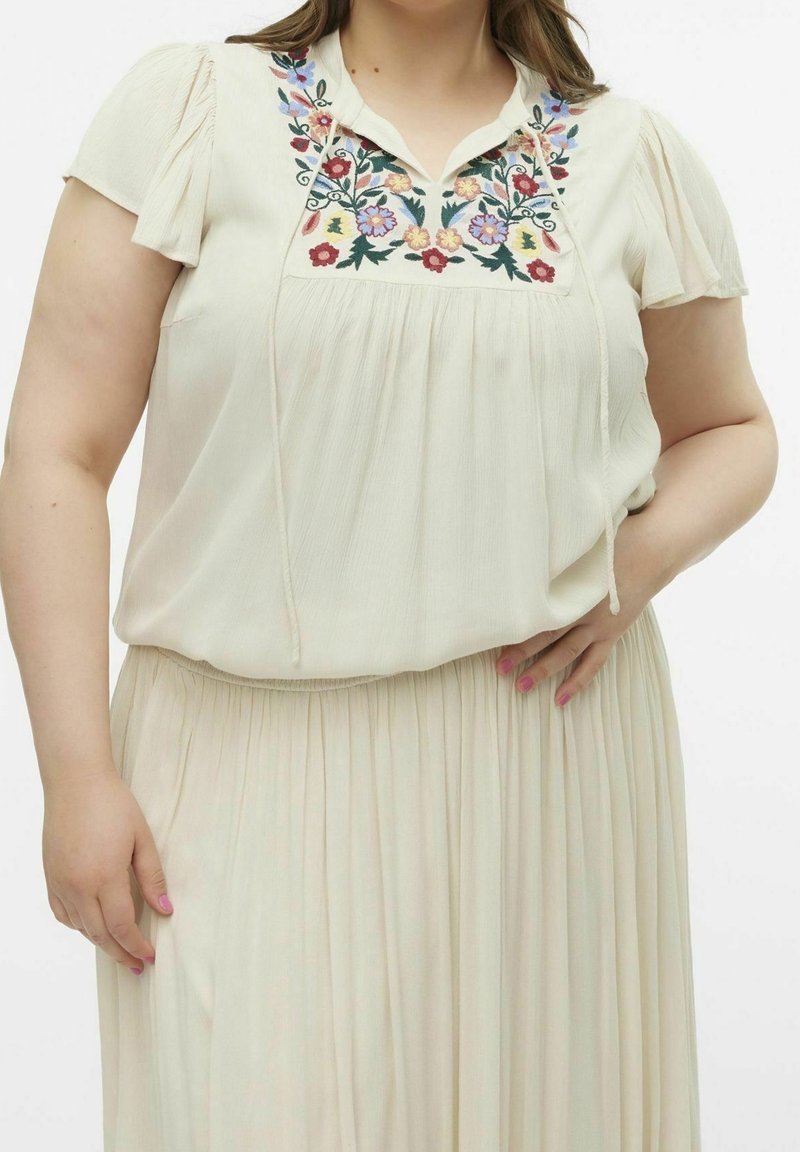Blouse de couleur crème avec broderies florales au niveau du col, en tissu doux, à manches courtes bouffantes et coupe ample, associée à une jupe plissée.