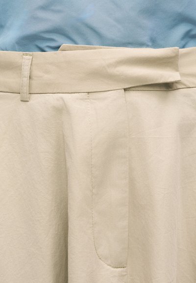 Béžové nohavice s vysokým pásom a širokým opaskom na modrom textilnom pozadí, zobrazujúce detailné prešívanie a textúru.