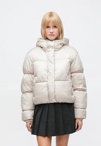 Lyshåret kvinde iført en lys beige dunjakke og en sort plisseret nederdel, vendt fremad mod en ensfarvet hvid baggrund.