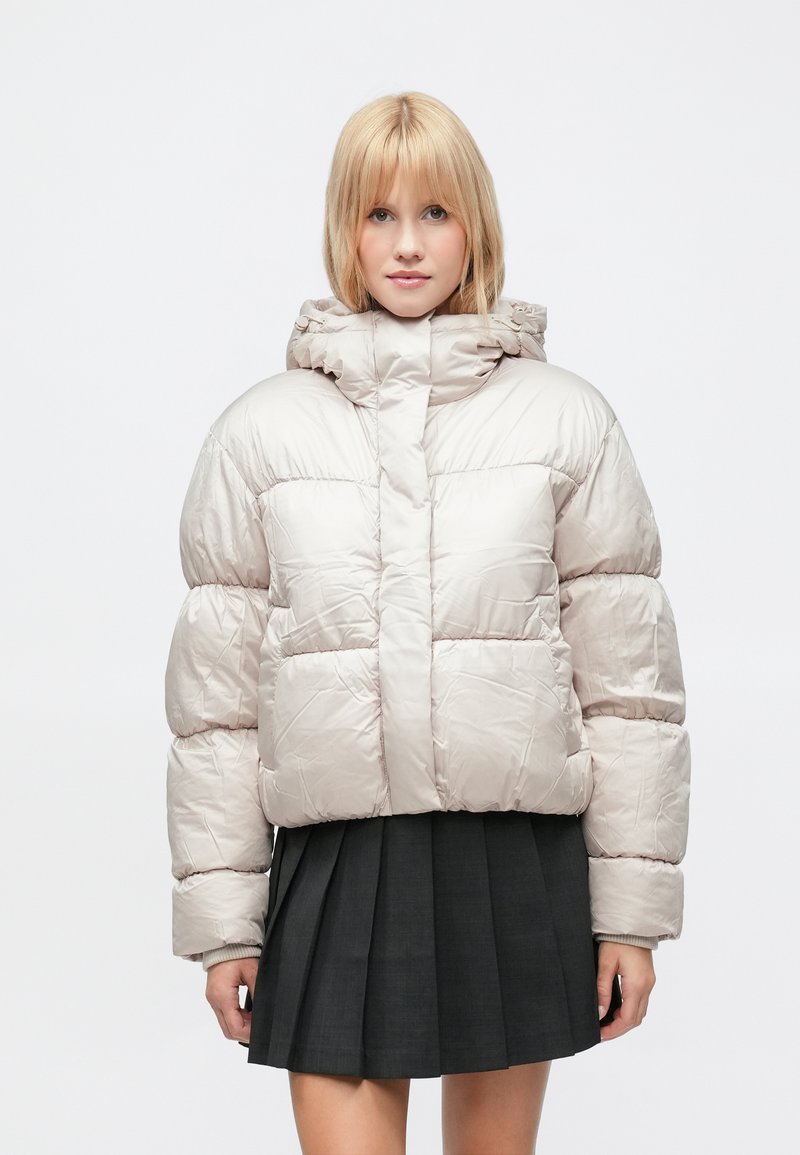 Lyshåret kvinde iført en lys beige dunjakke og en sort plisseret nederdel, vendt fremad mod en ensfarvet hvid baggrund.