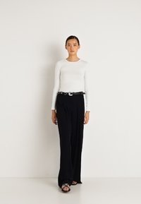 Egy nő áll egy fehér falnak támaszkodva, fehér hosszú ujjú felsőt, fekete, széles lábú nadrágot övvel és fekete szandált visel.