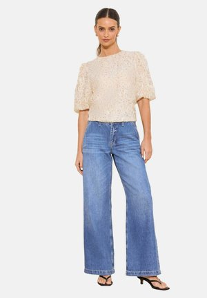 Vrouw staand, draag een crème kanten blouse met pofmouwen, blauwe hooggetailleerde wijde spijkerbroek en zwarte sandalen met hak tegen een witte achtergrond.