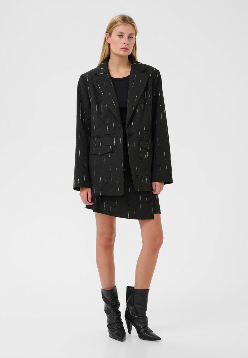 Blazer nero con una vestibilità su misura, tessuto strutturato, sequenze verticali e due tasche frontali, abbinato a una gonna coordinata e stivali al ginocchio.