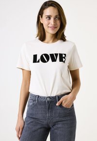Cremfarbendes Baumwoll-T-Shirt mit dem Aufdruck "LOVE" in fetten schwarzen Buchstaben. Kombiniert mit dunkelgrauen Jeans, die einen entspannten Schnitt und klassisches Design aufweisen.