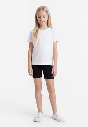 Jong meisje met lang blond haar, gekleed in een eenvoudig wit T-shirt, zwarte korte broek en witte sneakers, staand tegen een witte achtergrond.
