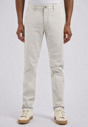 Man draagt licht beige rechte broek en gebroken witte sneakers, staand voor een effen lichte achtergrond.