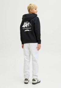 Čierna mikina s kapucňou má na chrbte grafikou hory a biely text; kombinovaná s svetlosivými teplákmi a čierno-bielymi teniskami.