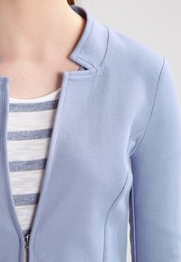 Ljusblå zip-jacka med rund krage, tillverkad av mjukt material, lager på lager över en vit och blå randig skjorta.