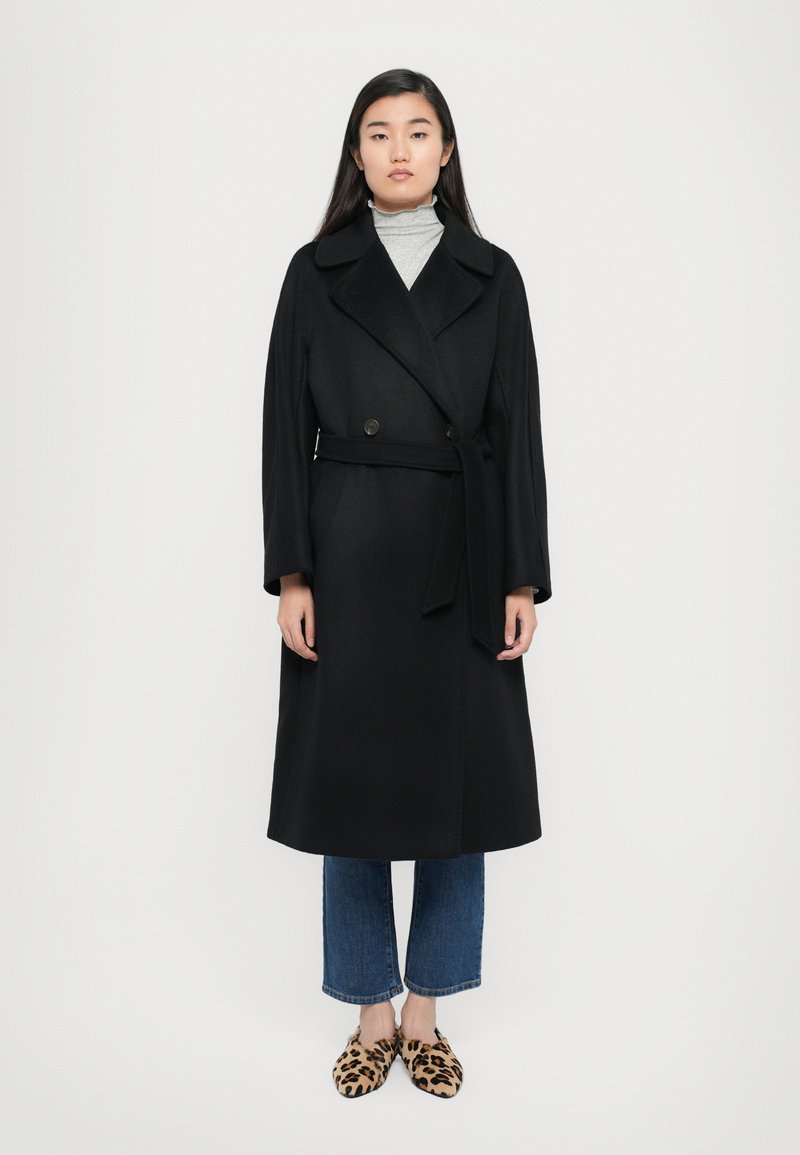Schwarzer Woll-Trenchcoat mit doppelreihigem Design, breiten Revers und einem Stoffgürtel, kombiniert mit kurz geschnittenen Jeans und Schuhen im Leopardenmuster.