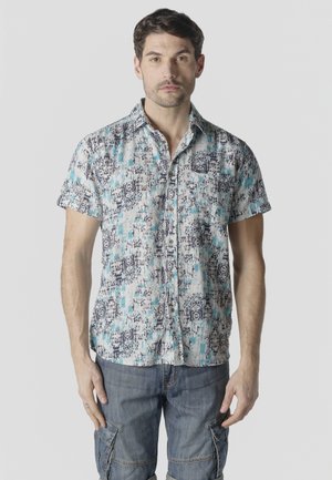 Hombre con una camisa de manga corta con botones, con un patrón abstracto azul y negro, y pantalones cortos cargo de mezclilla, frente a un fondo liso.