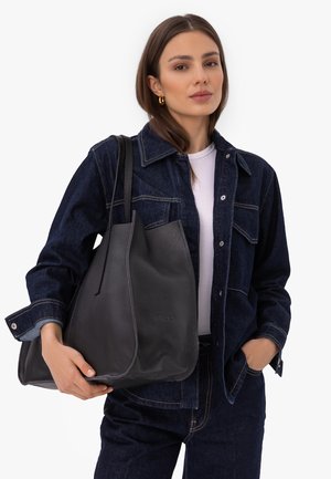 Joven mujer con chaqueta y pantalones de mezclilla oscuros sostiene un gran bolso tote de cuero negro sobre su hombro, de pie frente a un fondo blanco.