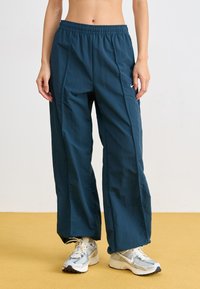 Τιρκουάζ, ελαφριά παντελόνια με φαρδιά πόδια, elastico στη μέση και πλευρικές ραφές. Συνδυασμένα με ασημένια αθλητικά παπούτσια σε κίτρινο δάπεδο.