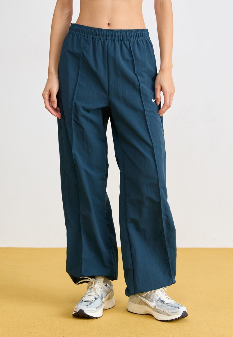 Τιρκουάζ, ελαφριά παντελόνια με φαρδιά πόδια, elastico στη μέση και πλευρικές ραφές. Συνδυασμένα με ασημένια αθλητικά παπούτσια σε κίτρινο δάπεδο.