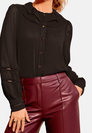 Blouse noire à manches longues avec boutons en forme de cœur rentrée dans un pantalon en cuir bordeaux taille haute, modèle avec la main dans la poche.