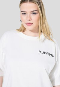 Camiseta blanca de gran tamaño hecha de algodón, con "PALM SPRINGS" bordado en negrita de color negro en el área del pecho izquierdo. Cuello redondo, mangas cortas.