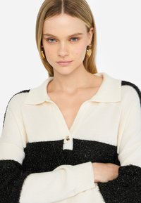 Maglione polo lavorato a maglia con corpo color crema e una fascia orizzontale soffice nera, chiusura con bottoni dorati e maniche testurizzate a contrasto.