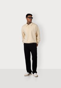 Beige Polo-Sweatshirt mit lockerer Passform, schwarze Hose und weiße Sneakers. Das Polo hat einen dezenten Kragen und ein gesticktes Logodetail.