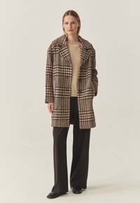 Bruine en crèmekleurige geblokte jas met een oversized kraag, knopen aan de voorkant en zakken, gecombineerd met een beige sweater en zwarte wijde broek.