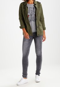 Veste militaire verte, t-shirt graphique gris avec texte et impression de rose, jean skinny gris, et baskets blanches.