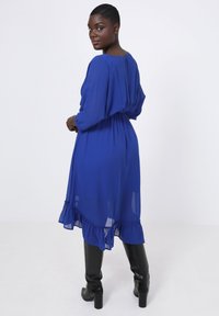 Blauwe, vloeiende jurk met lange, opgezette mouwen, elastische tailleband, gerimpelde zoom, gemaakt van lichte stof, gecombineerd met zwarte knieën hoge laarzen.