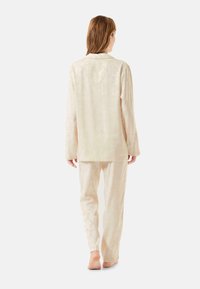 Conjunto de pijama en suave tela color crema claro con un sutil patrón floral. La parte superior tiene cuello y mangas largas; los pantalones son de corte suelto.