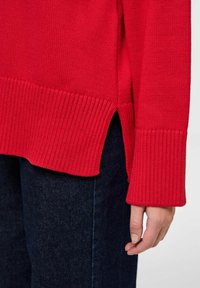 Czerwony sweter z dzianiny z prążkowanym dołem i mankietami, z bocznymi rozcięciami. Noszony na ciemne dżinsy, podkreślający fakturę i żywy kontrast kolorów.