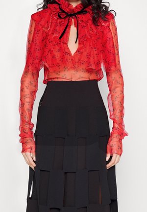 Blouse rouge transparente à motifs floraux, col à volants et nœud noir ; associée à une jupe noire à volants avec découpes rectangulaires.