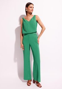 Zielony kombinezon bez rękawów z dekoltem w kształcie litery V i ramiączkami, z szerokimi spodniami. W zestawieniu z brązowymi sandałami na słupku.