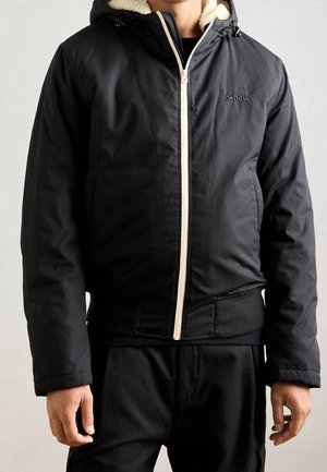 Homme portant une veste noire zippée avec une capuche doublée en polaire crème et un pantalon noir, debout devant un fond clair uni.