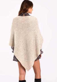 Poncho de punto beige con forma triangular, que presenta una textura suave y sutiles motas, llevado sobre un vestido floral oscuro.